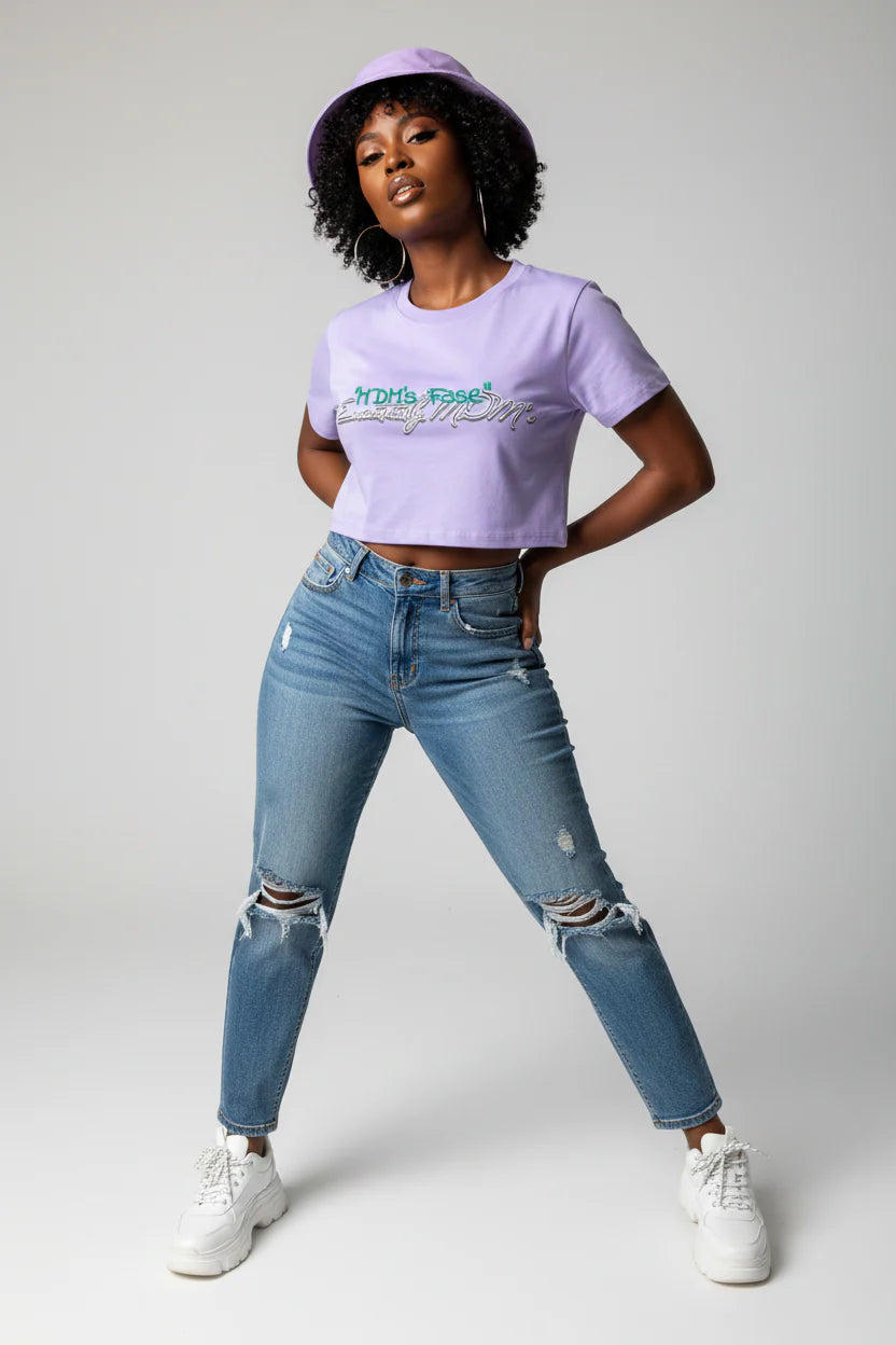 A model posing in a studio wearing a light purple cropped t-shirt with a graphic print, blue distressed jeans, white sneakers, and a matching purple bucket hat.